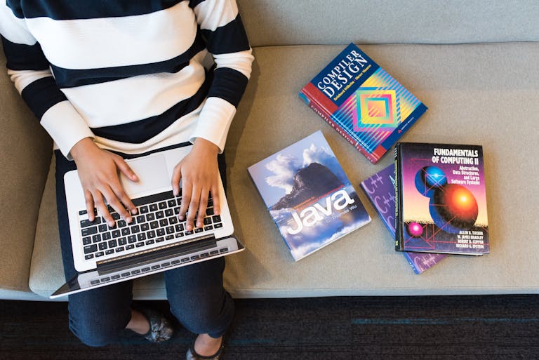 Woman using laptop on sofa, surrounded by programming books, learning coding.